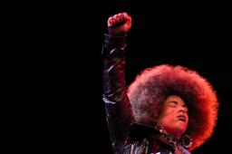 Brown-skinned woman with large natural afro holds fist in air. She is wearing a reflective black trench coat. Her eyes are closed and her face tilts up toward blush pink lights.